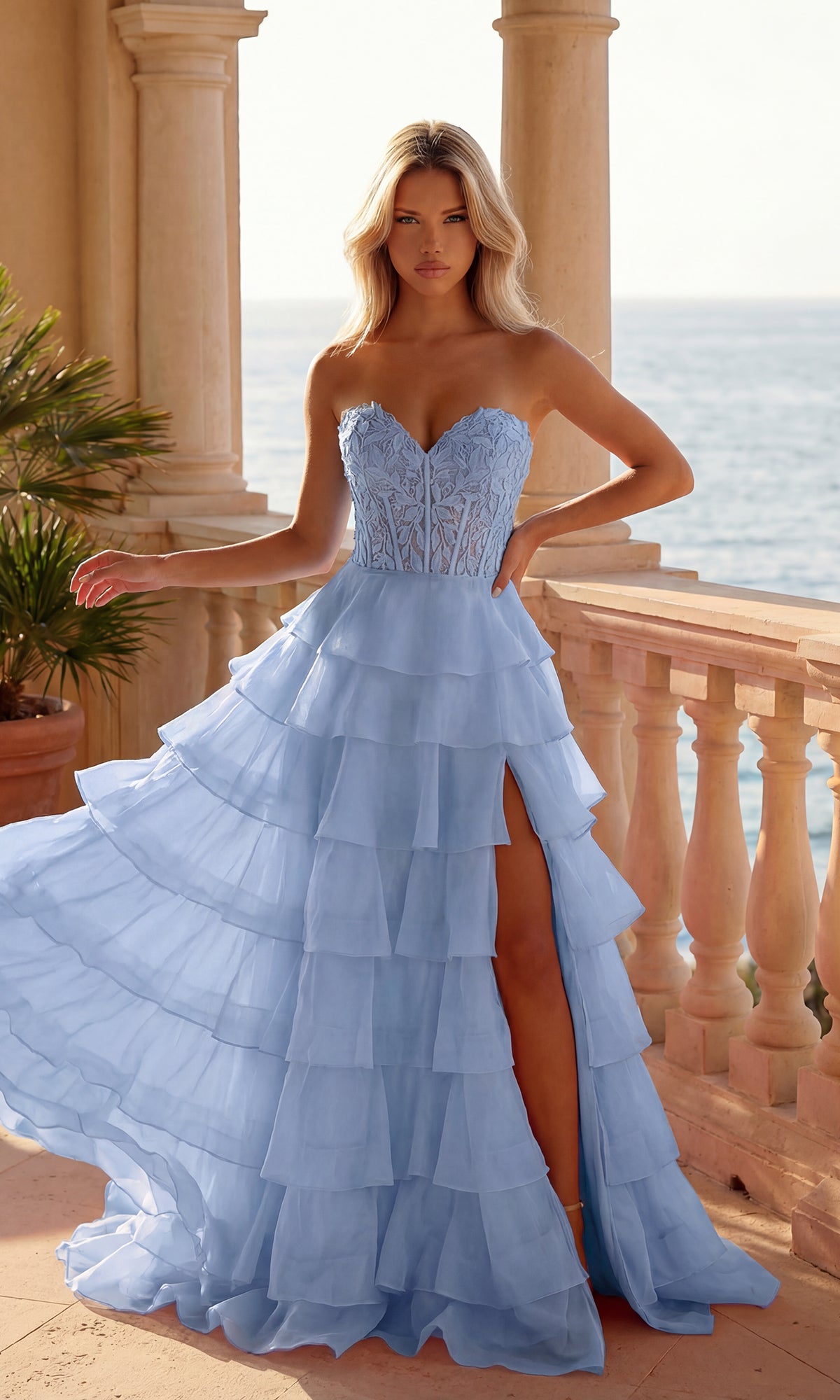 A woman in a periwinkle Strapless Long Ruffled Prom Gown (PromGirl 250001) with a sheer corset-style bodice and thigh-high slit stands on a balcony overlooking the ocean.